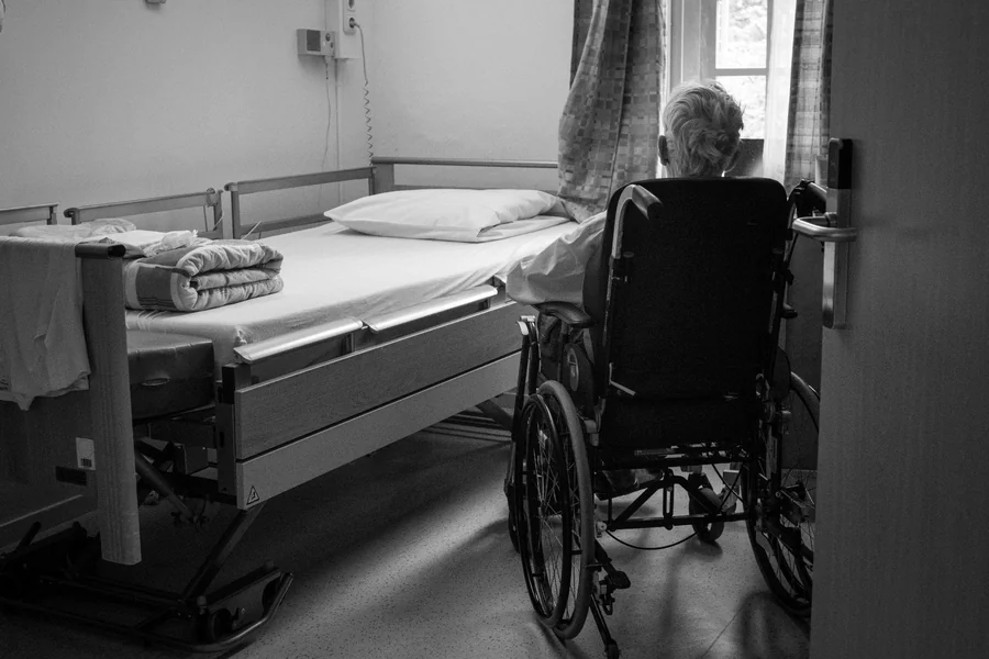 Elderly nursing home resident in a wheelchair beside a hospital bed, highlighting fall risk and neglect concerns in long-term care facilities
